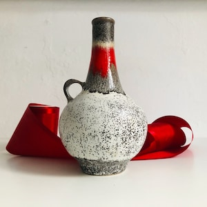 Könnte beinhalten: Eine weiße Keramikvase mit gesprenkeltem Überzug und einem roten und grauen Band um den Hals. Die Vase hat einen runden Körper und einen schmalen Hals mit einem kleinen Griff.