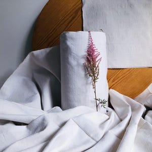 以下が含まれることがあります： 白いリネン生地のロールに、ピンクの花の茎が置かれています。生地は木製の表面にかけられています。