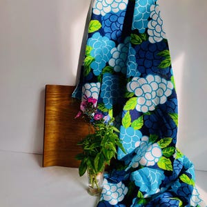 Puede incluir: Una tela drapeada con un estampado floral en tonos azules y blancos, acentuada con hojas verdes. La tela está drapeada sobre una tabla de madera y junto a un vaso con plantas y flores verdes.