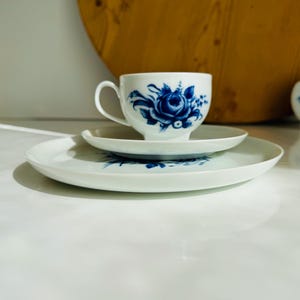 May include: A white porcelain teacup, saucer, and plate set with a blue floral design. The teacup sits atop the saucer, which is on a larger plate. A small jar with a matching design is in the background. The set is on a white surface.