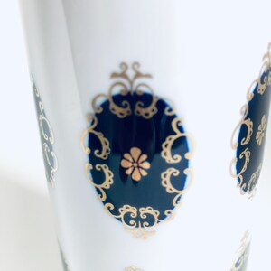 Könnte beinhalten: Weißer Keramikbecher mit einem sich wiederholenden Muster aus dekorativen Medaillons in Marineblau und Gold. Jedes Medaillon hat einen marineblauen Hintergrund mit einem goldenen Blumendesign in der Mitte und goldenen Filigranarbeiten an den Rändern.