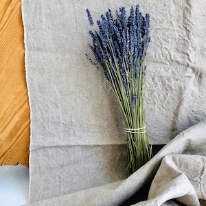 Könnte beinhalten: Ein Bündel getrockneter Lavendelblüten mit leuchtend violetten Blüten und grünen Stielen, gebunden mit einem Naturgummiband. Der Lavendel liegt auf einem strukturierten, hellgrauen Leinentuch, mit einem Holzuntergrund im Hintergrund.