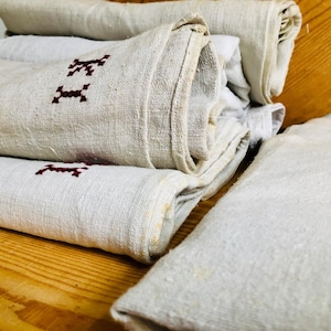 Op de afbeelding: Een stapel witte linnen theedoeken met rode geborduurde letters. De handdoeken zijn opgerold en gestapeld op een houten oppervlak.