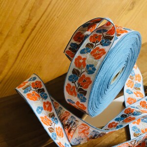 Può includere: Un rotolo di nastro blu e bianco con un motivo floreale in arancione, marrone e blu. Il nastro è rifinito con un bordo bianco.