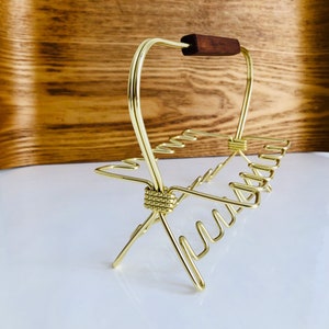 Könnte beinhalten: Ein goldfarbenes Metall-Serviertablett mit einem dunkelbraunen Holzgriff. Das Tablett hat ein einzigartiges, offenes Design mit mehreren gebogenen Haltern. Der Griff ist rechteckig und scheint aus Holz zu sein.