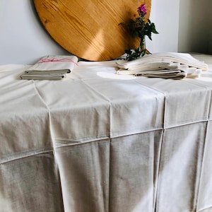 Könnte beinhalten: Eine cremefarbene Tischdecke bedeckt einen Tisch mit gefalteten Servietten und einem Stapel gefalteter Bettwäsche. Im Hintergrund befindet sich ein rundes Holzobjekt sowie eine kleine Vase mit Blumen. Die Bettwäsche ist mit rosa Schnur gebunden.