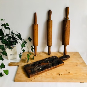 Può includere: Tre antichi mattarelli in legno di varie dimensioni e uno stampo in legno intagliato poggiano su un grande tagliere in legno. I mattarelli sono di colore marrone caldo e lo stampo è marrone scuro. Sullo sfondo c'è una piccola pianta in un vaso bianco.