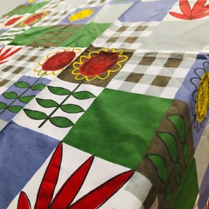 May include: A white tablecloth with a checkered pattern in brown and white. The tablecloth has a patchwork design with green, blue, and white squares. Each square features a different floral design in red, yellow, and green.