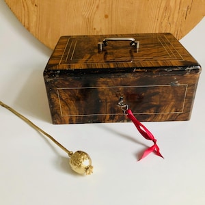 Könnte beinhalten: Eine Vintage-Holzkiste mit Schlüssel und rotem Band sowie einem goldfarbenen dekorativen Stiel mit einer Samenkapsel. Die Kiste hat ein dunkles Holzmaserungsmuster und einen Metallgriff. Der Schlüssel steckt im Schloss.