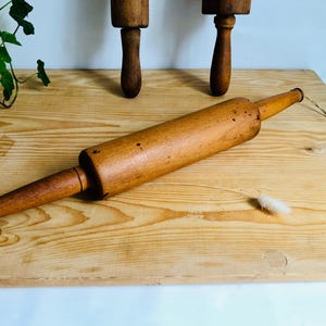 Può includere: Un mattarello di legno con estremità affusolate e corpo cilindrico è appoggiato su una superficie di legno chiaro. Sullo sfondo sono visibili altri due mattarelli. Il mattarello è un utensile da cucina per appiattire l'impasto.
