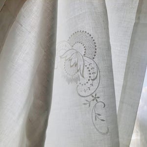 Puede incluir: Un panel de cortina blanco semitransparente con un delicado motivo floral bordado en tonos claros. La tela tiene una textura tejida visible y el bordado incluye formas complejas de pétalos, líneas curvas y pequeños puntos sutiles.