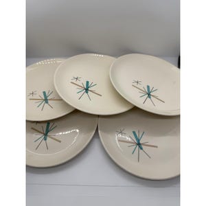 May include: A set of four off-white ceramic plates, each featuring a central graphic pattern of intersecting lines in black, teal, and tan. The design consists of thin radiating spokes and a solid rectangular bar, creating a starburst motif.