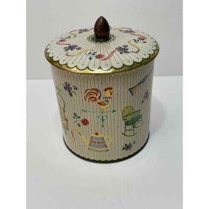 May include: A cylindrical metal canister with a cream-colored background and vertical red pinstripes. The container features a removable lid with a dark brown knob and is decorated with colorful illustrations of a rooster, a rocking chair, a clock, and flowers.