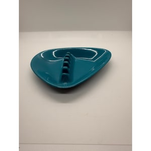 May include: A teal-colored ceramic ashtray with a smooth, glossy finish and an asymmetrical, triangular shape. The center features a raised, integrated bridge with five distinct notches for resting items, set against a solid background.