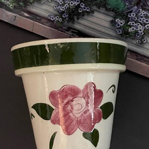 Puede incluir: Una maceta de cerámica blanca con borde verde y diseño de flor rosa pintada a mano. La maceta tiene una forma clásica y es adecuada para plantas pequeñas o hierbas. La maceta mide aproximadamente 10 cm de altura.