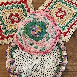 Op de afbeelding: Een verzameling van vier handgemaakte haakwerkjes. De items omvatten twee vierkante pannenlappen met rode, groene en gele patronen, een ronde roze en groene pannenlap en een witte doily met een gerimpelde rand. De items worden gepresenteerd op een houten oppervlak.