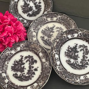 May include: A set of four small, decorative plates with a black and white Asian-inspired design. Each plate features a central landscape scene surrounded by a detailed border. The plates are arranged with a vibrant pink flower arrangement in the background.