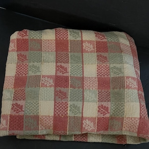 May include: A folded fabric textile featuring a checkered pattern in shades of muted red, sage green, and cream. The design includes alternating squares with a woven leaf motif and a textured grid pattern, creating a consistent repeating visual layout.
