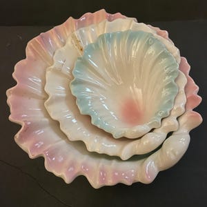 Puede incluir: Un juego de tres platos de cerámica con forma de concha de diferentes tamaños. El plato más grande tiene un borde con volantes con tonos rosas y blancos, mientras que los platos más pequeños presentan acentos en azul pastel y rosa. Los platos están apilados.