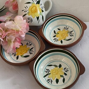 May include: A white ceramic pitcher and three matching bowls. The pitcher and bowls feature a yellow floral design with green leaves. The bowls have brown rims and blue and green stripes. Pink flowers are in the background.