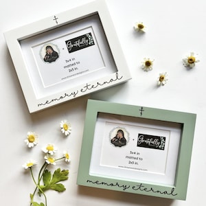 Puede incluir: Dos marcos de fotos blancos y verdes con un diseño de cruz. Los marcos miden 7,6 x 10,2 cm y están enmatados a 5,1 x 7,6 cm. Los marcos tienen el texto "memory eternal" y "Gratefully Designed".