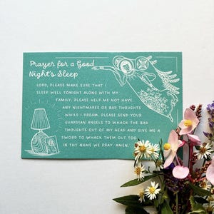 Puede incluir: Una tarjeta turquesa con el texto "Prayer for a Good Night's Sleep" y una oración. La tarjeta presenta un dibujo lineal de un ángel y una lámpara. Flores con pétalos rosas y blancos están en el fondo.