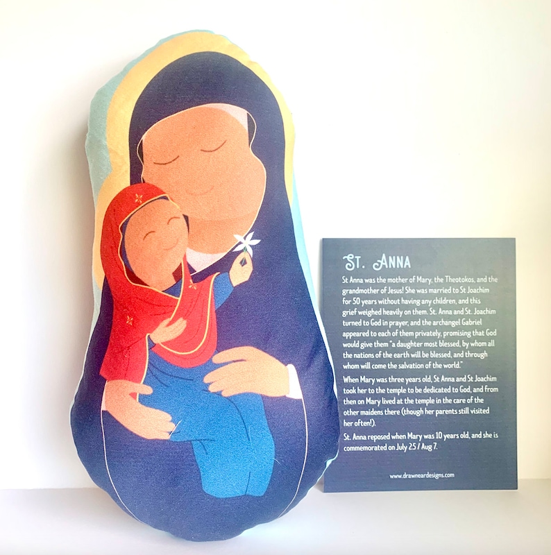 May include: A blue and red plush pillow with an illustration of Saint Anne holding the baby Jesus. The pillow is shaped like a teardrop and has a gold halo around the figures. The text on the card reads "St. Anna".