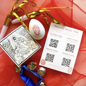 May include: An Easter-themed collection featuring a white egg with "Christ is Risen" text, a sticker with religious imagery, a blue tambourine with bells, and a card with QR codes for Pascha music.