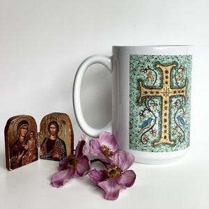 May include: A white ceramic mug with a large handle, featuring a mosaic-style cross design in gold and green. Two small religious icons and purple flowers are also present.