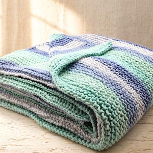 May include: A folded, knitted blanket in shades of blue, teal, and white. The blanket is made of soft yarn and has a textured appearance. The blanket is folded neatly, with a corner turned over, and is suitable for home decor.