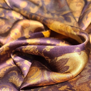 May include: Close-up of a silky fabric with a gold and purple floral pattern. The fabric is folded and draped, showing the texture and sheen of the material.