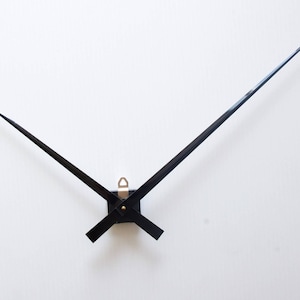 Op de afbeelding: Zwarte wijzers van een klok met een zilveren middelpunt. De wijzers zijn lang en dun en zijn geplaatst tegen een witte achtergrond.