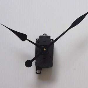 Könnte beinhalten: Schwarze Uhrzeiger, die an einem schwarzen Uhrwerk befestigt sind. Die Zeiger befinden sich in Kreuzstellung.