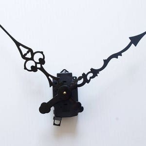 May include: Black clock hands with a decorative design. The hands are attached to a black clock mechanism.