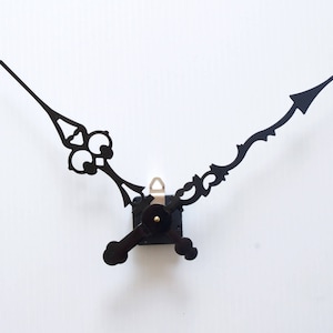 Op de afbeelding: Zwarte wijzers met een decoratief ontwerp. De wijzers zijn bevestigd aan een zwart uurwerk.