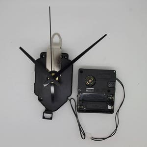 Può includere: Meccanismo di orologio nero con lancette e un pacco batteria. Il meccanismo dell'orologio ha un gambo in metallo argentato e un alloggiamento in plastica nera. Il pacco batteria è nero e ha un'etichetta che dice "1.5V DC".