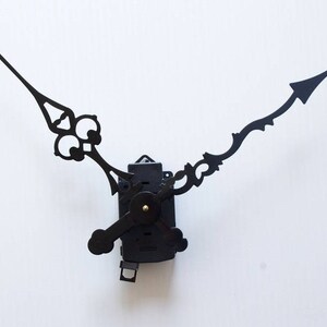 May include: Black clock hands with a decorative design. The hands are attached to a black clock mechanism.