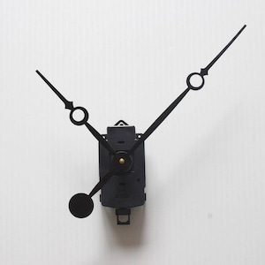 May include: Black clock movement with hour, minute, and second hands. The hands are thin and pointed, with circular accents. The clock mechanism is rectangular with a small pendulum and a brass-colored center.