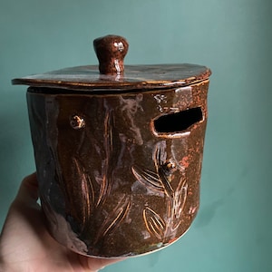 Könnte beinhalten: Eine braune Keramikdose mit Deckel und floralem Muster. Die Dose hat eine kleine Öffnung an der Seite.