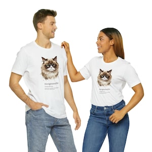 Könnte beinhalten: Zwei weiße T-Shirts mit einer Illustration eines mürrischen Katers und dem Wort "morgenmuffel" darauf gedruckt. Das Wort ist definiert als "eine Person, die morgens schlecht gelaunt ist".