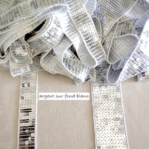 Op de afbeelding: Zilveren paillettenband met een witte rand, losjes gerangschikt. De band is gemaakt van kleine, vierkante pailletten die licht reflecteren. De tekst "argent sur fond blanc" is zichtbaar.