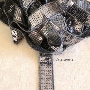 Op de afbeelding: Een rol grijze paillettenbies met een metallic glans. De bies is gemaakt van kleine, vierkante pailletten die op een stoffen achterkant zijn genaaid. De tekst "Gris souris" is zichtbaar.