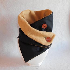 Puede incluir: Un calentador de cuello de vell&oacute;n negro y marr&oacute;n con dos botones de madera. El calentador est&aacute; doblado por la mitad y se muestra en un soporte blanco.
