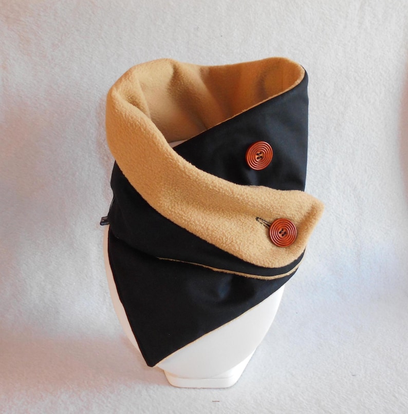 Puede incluir: Un calentador de cuello de vell&oacute;n negro y marr&oacute;n con dos botones de madera. El calentador est&aacute; doblado por la mitad y se muestra en un soporte blanco.