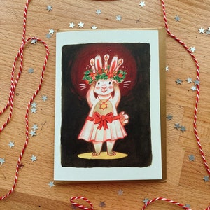 May include: A greeting card featuring a whimsical illustration of a rabbit wearing a candle-lit crown and a dress with a red bow. The card is set against a dark red background, with a brown envelope. Red and white twine and star-shaped confetti surround the card on a wooden surface.