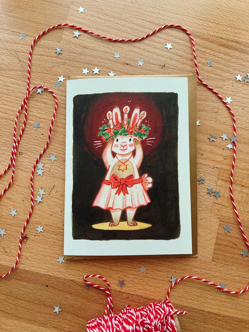 May include: A greeting card featuring a whimsical illustration of a rabbit wearing a candle-lit crown and a dress with a red bow. The card is set against a dark red background, with a brown envelope. Red and white twine and star-shaped confetti surround the card on a wooden surface.