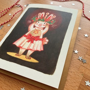 May include: A Christmas card featuring a cartoon rabbit wearing a festive wreath and dress, with a red bow. The rabbit is on a stage with a spotlight. The card is on a brown envelope, with red and white twine and star-shaped confetti.