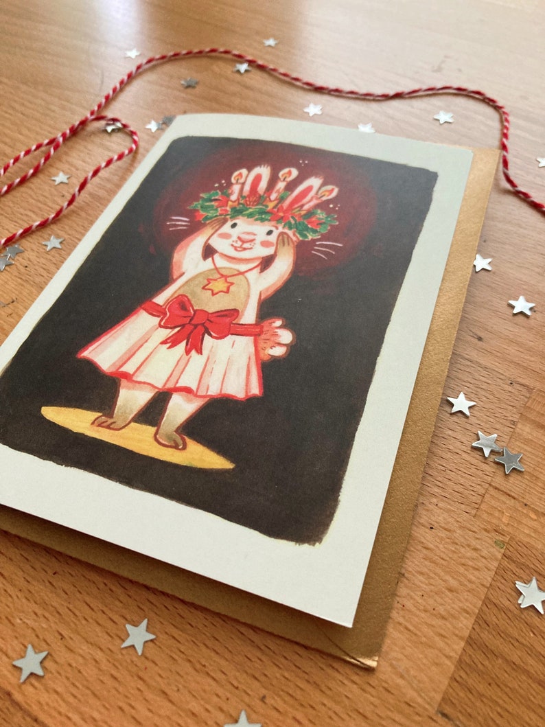 May include: A Christmas card featuring a cartoon rabbit wearing a festive wreath and dress, with a red bow. The rabbit is on a stage with a spotlight. The card is on a brown envelope, with red and white twine and star-shaped confetti.
