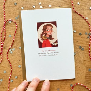 May include: A Christmas card featuring an illustration of a woman with a bird on her shoulder. The card has the text "Christmas Carol 'St Lucia'" and is decorated with red and white twine and silver star-shaped confetti.