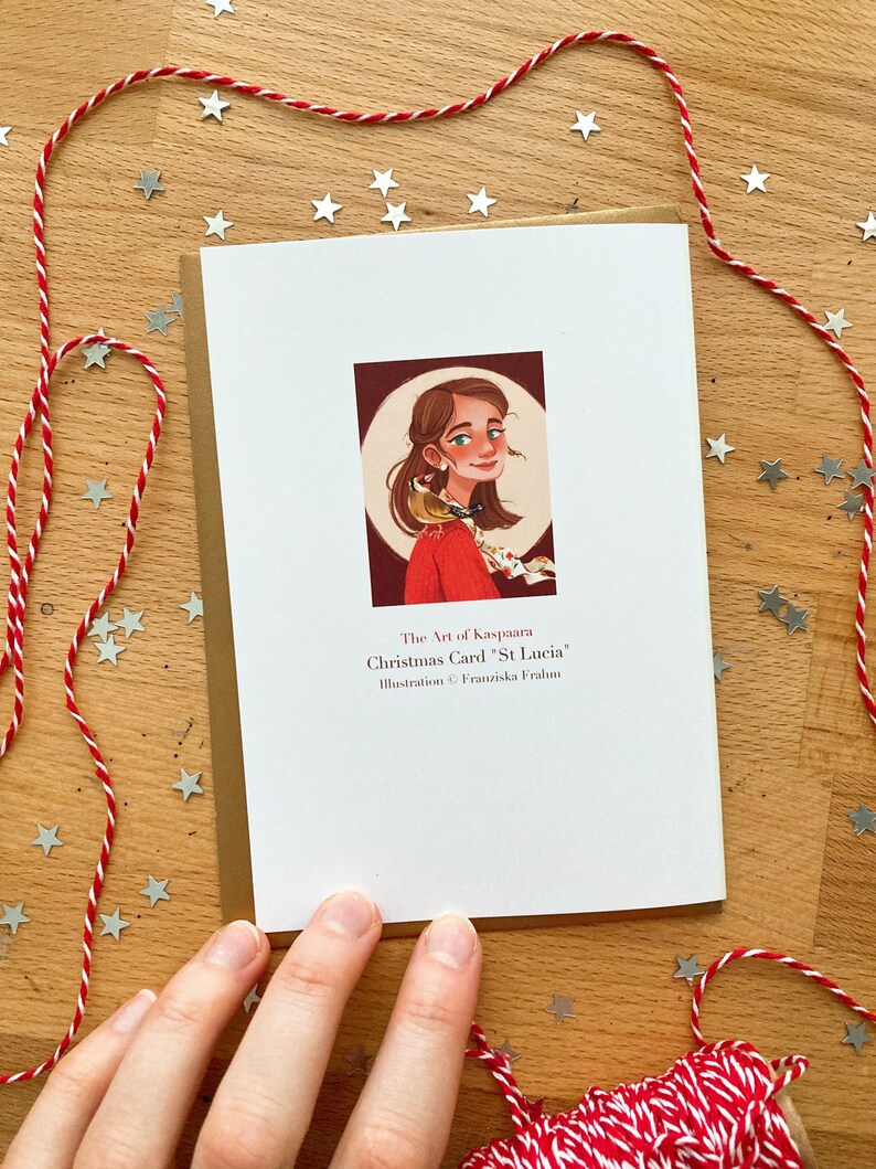 May include: A Christmas card featuring an illustration of a woman with a bird on her shoulder. The card has the text "Christmas Carol 'St Lucia'" and is decorated with red and white twine and silver star-shaped confetti.